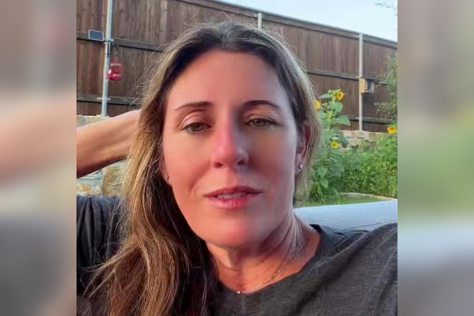 Sheila Greene sits outside on her back patio in a dark tee shirt with one hand behind her head, talking to the camera. Behind her are a wooden privacy fence, a red hummingbird feeder, tall sunflowers, and green garden plants in warm late day light.