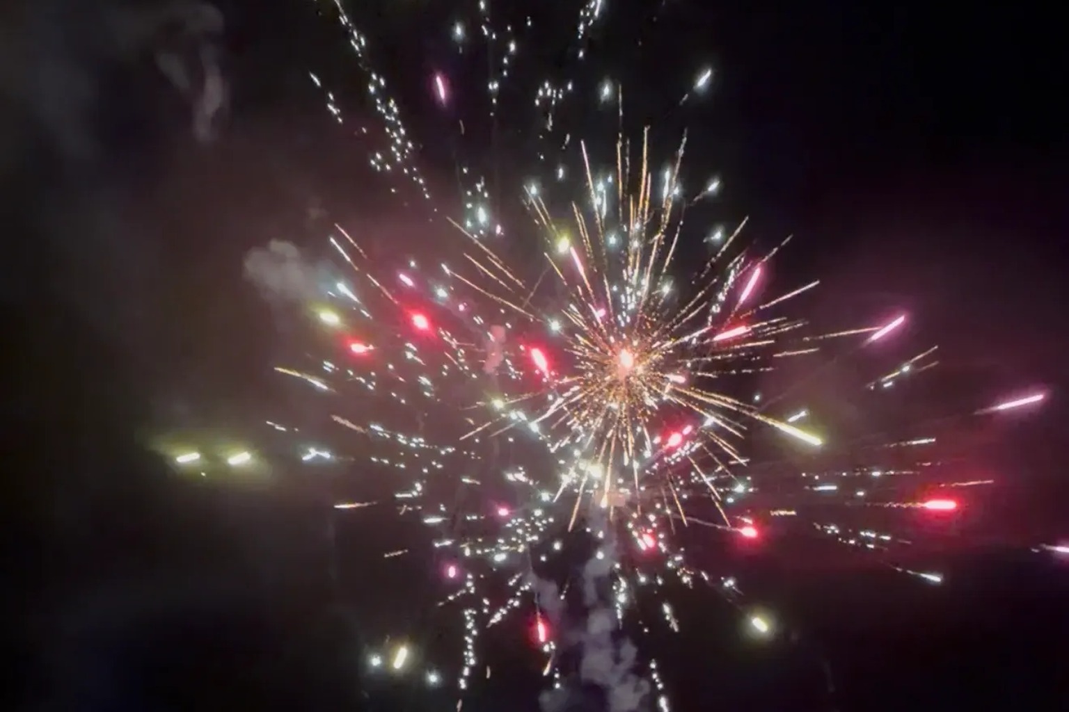 Colorful fireworks burst against a dark night sky, with red, gold, pink and white sparks radiating outward and a smoke trail rising from below.