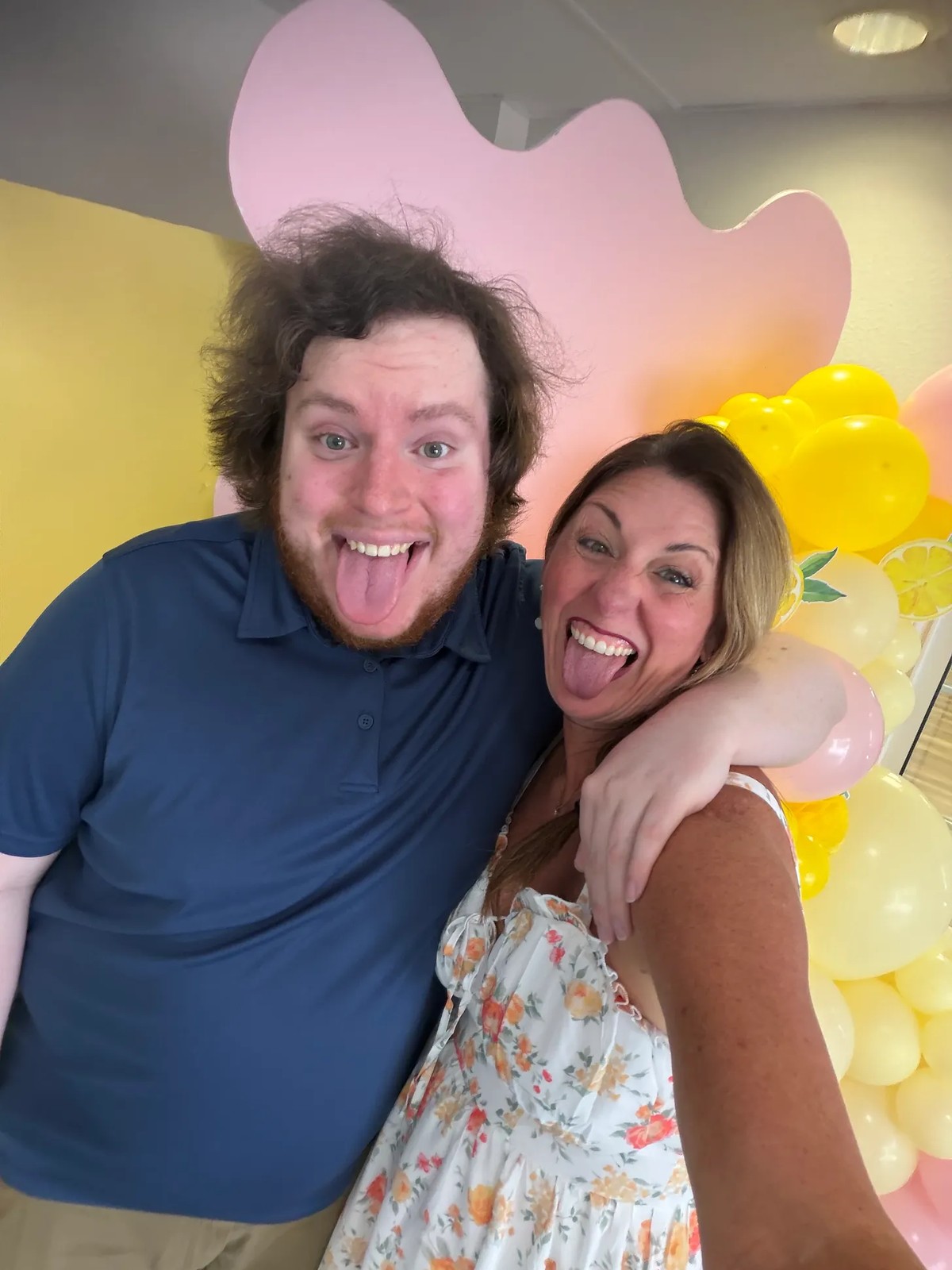 Sheila and Nathan leaning in for a selfie in front of the pink and yellow balloon arch, both sticking their tongues out and laughing.