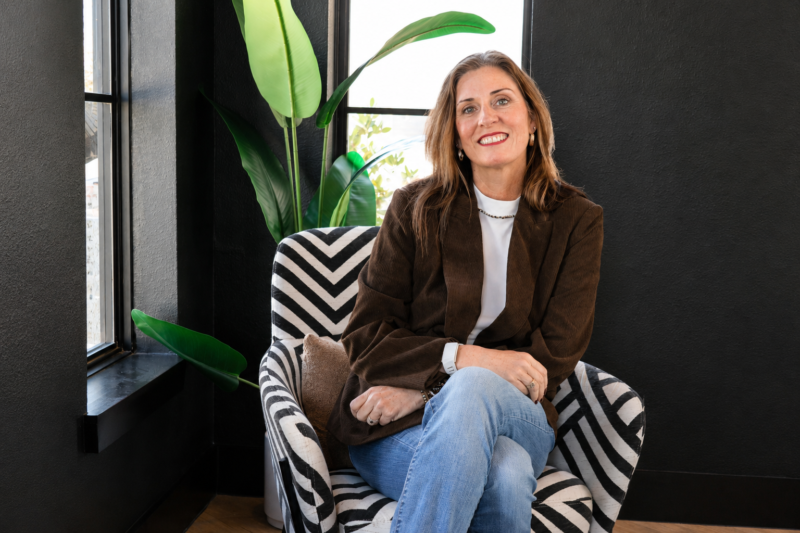 Sheila Greene seated in a patterned armchair beside a tall green plant, smiling.